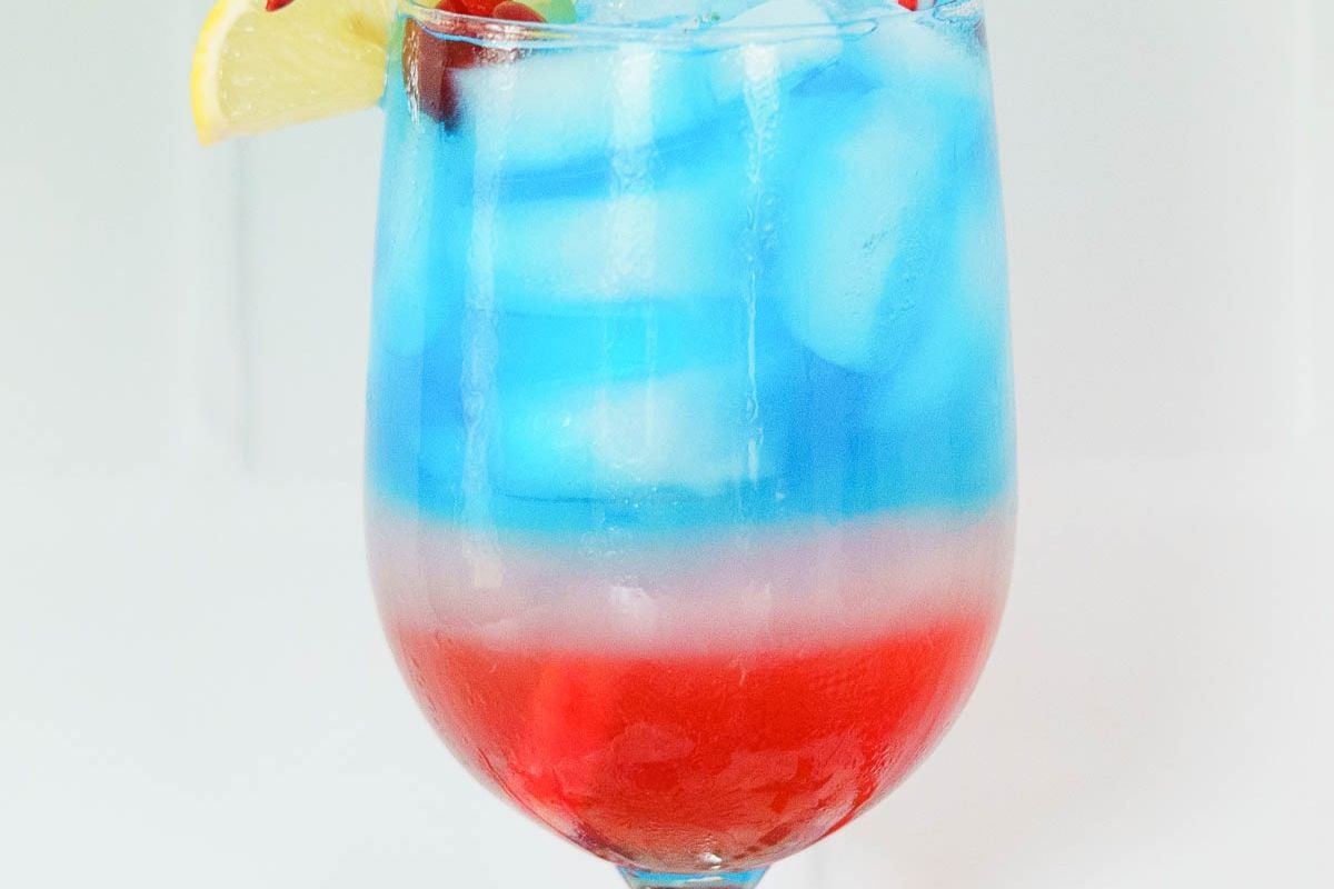 A glass with a layered red, white, and blue drink—perfect for Labor Day—topped with ice, a lemon wedge, and a cherry, set against a plain white background. This festive beverage is the ideal way to cool off in style.