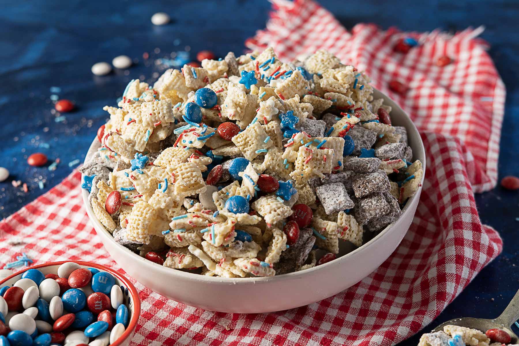 A bowl of Chex mix with red, white, and blue candies, sprinkles, and cereal pieces sits on a red plaid cloth with scattered candies around—perfect for cookouts or as an easy dessert for your Labor Day celebration.