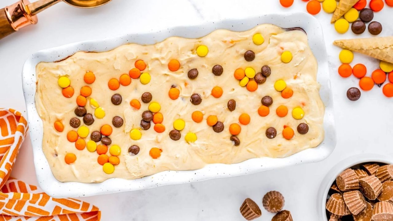 A rectangular dish of ice cream topped with orange, yellow, and brown candy-coated chocolates, surrounded by similar candies and mini peanut butter cups on a white surface—perfect for fans of peanut butter desserts looking to try new dessert recipes.