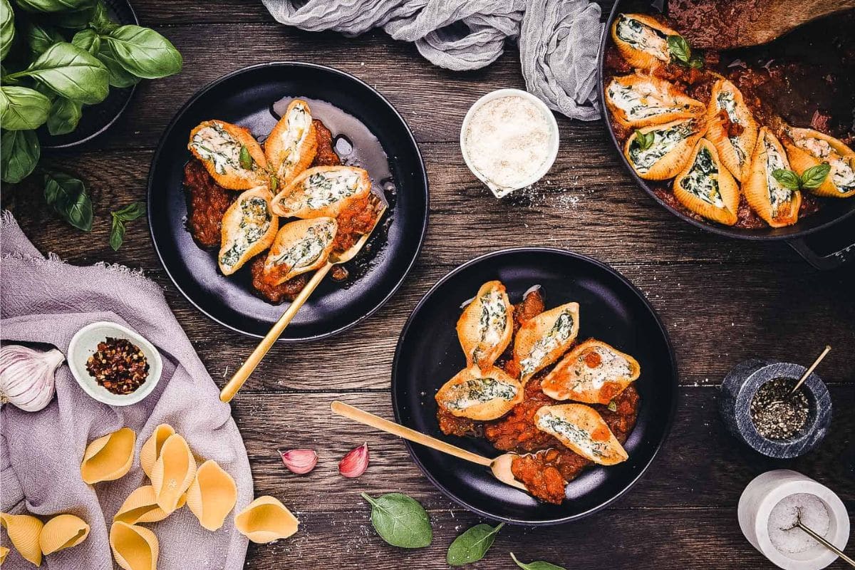 Three black bowls of stuffed pasta shells with tomato sauce sit on a dark table, surrounded by loose pasta, basil leaves, garlic, and seasonings—a perfect scene for cozy meals and comforting pasta dinners.
