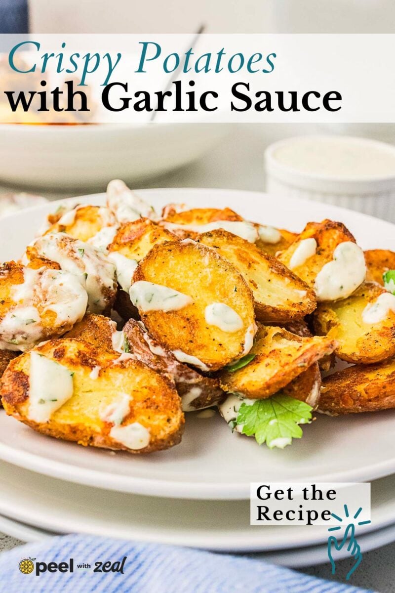 A plate of crispy roasted red skin potato halves topped with creamy garlic sauce, garnished with parsley, with a small bowl of sauce in the background.