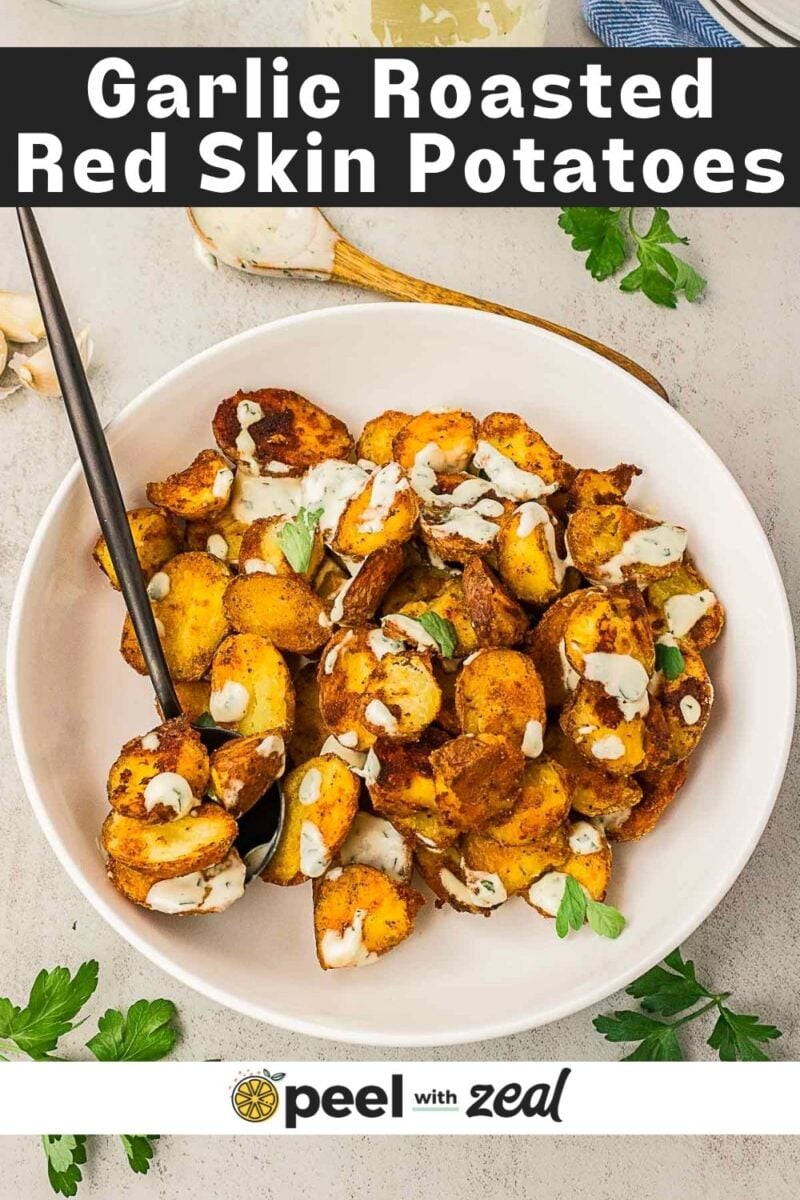 Bowl of perfectly roasted red skin potatoes topped with creamy sauce and herbs, served with a spoon, on a white surface with parsley scattered around.