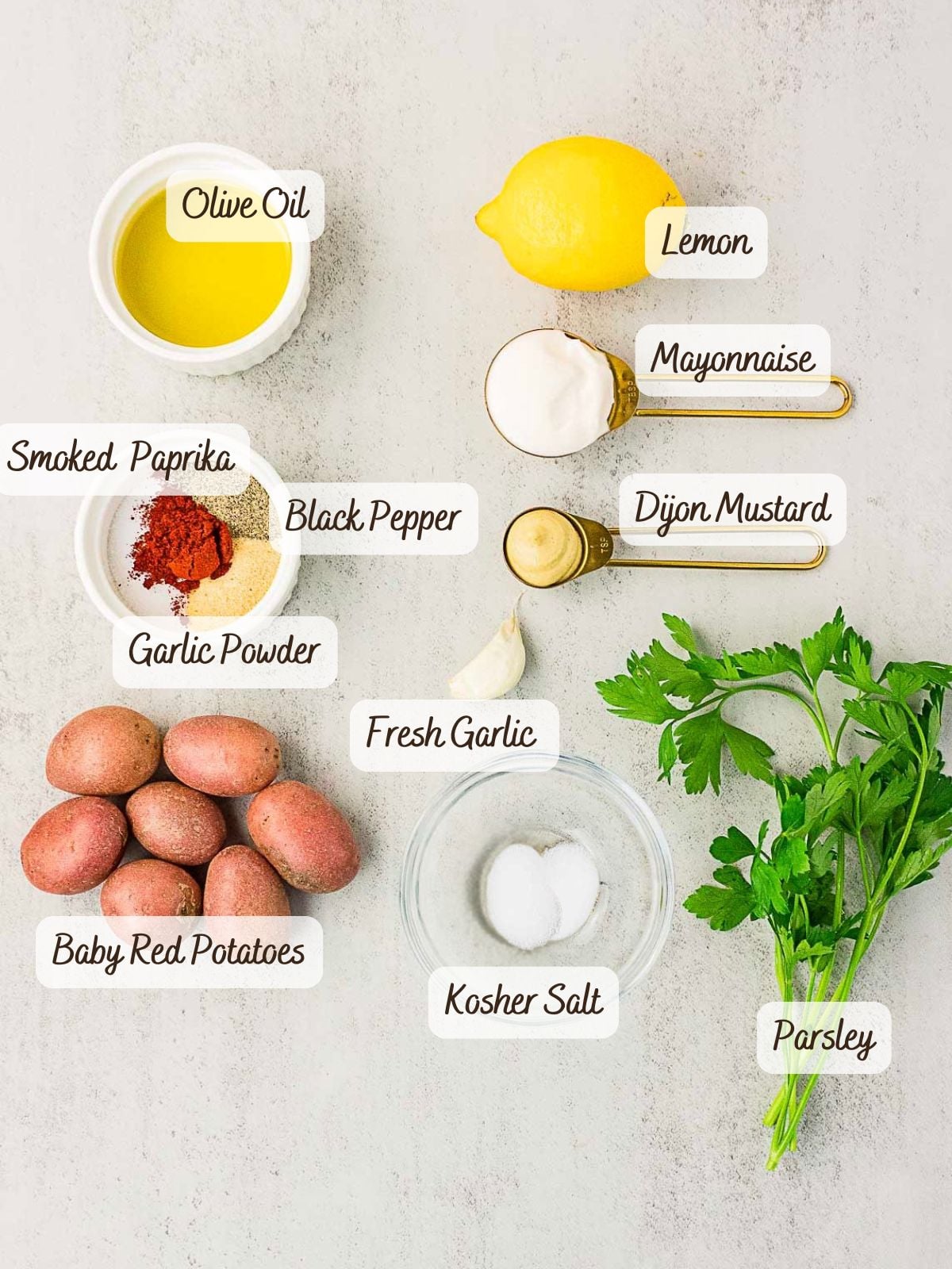 Ingredients arranged on a white surface for roasted red skin potatoes: olive oil, lemon, mayonnaise, Dijon mustard, smoked paprika, black pepper, garlic powder, fresh garlic, baby red potatoes, kosher salt, and parsley.