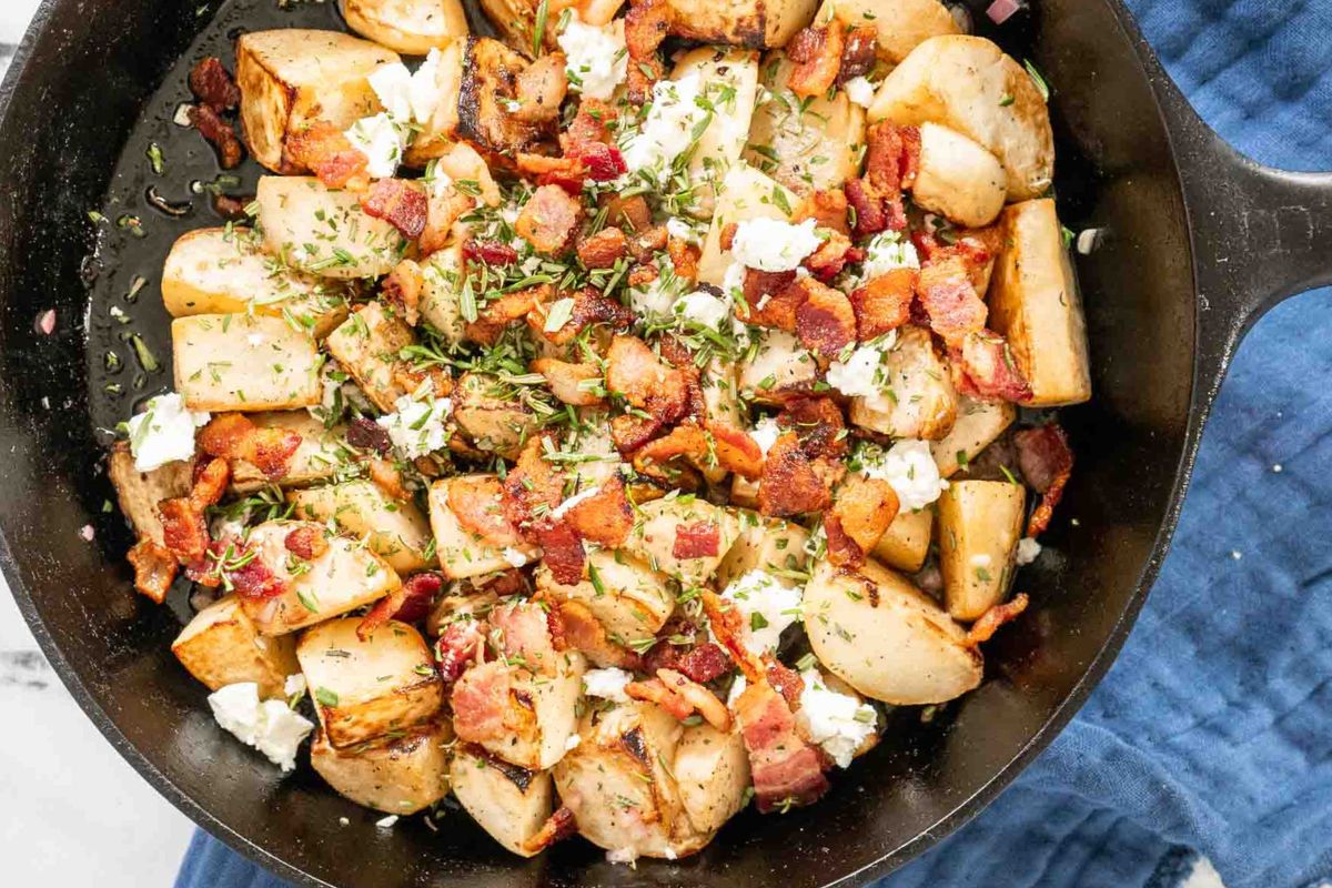 Cast iron skillet filled with roasted potatoes, crumbled bacon, goat cheese, and chopped herbs on a blue cloth background—a comforting addition to your collection of vegetable recipes that highlights the delicious possibilities of cooking with vegetables.