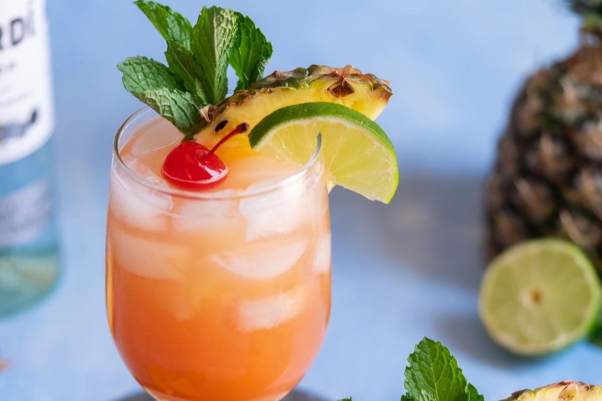 A tropical cocktail with ice, garnished with a pineapple slice, lime wheel, cherry, and mint sprig—perfect to cool off and enjoy refreshing beverages. A pineapple and lime are visible in the background.