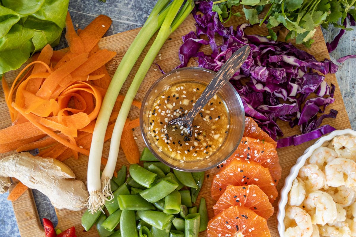 A cutting board with carrots, green onions, ginger, snap peas, red cabbage, cilantro, orange slices, shrimp, and a bowl of homemade salad dressing creates the perfect setup for fresh homemade recipes.