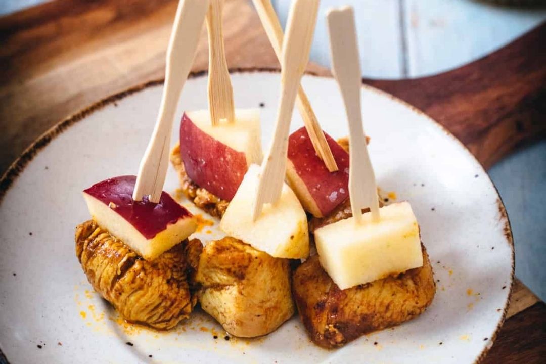 A white plate holds pieces of cooked chicken topped with apple cubes, each skewered with a small wooden pick—a simple yet elegant dish that’s perfect for experimenting with apple recipes during apple season.