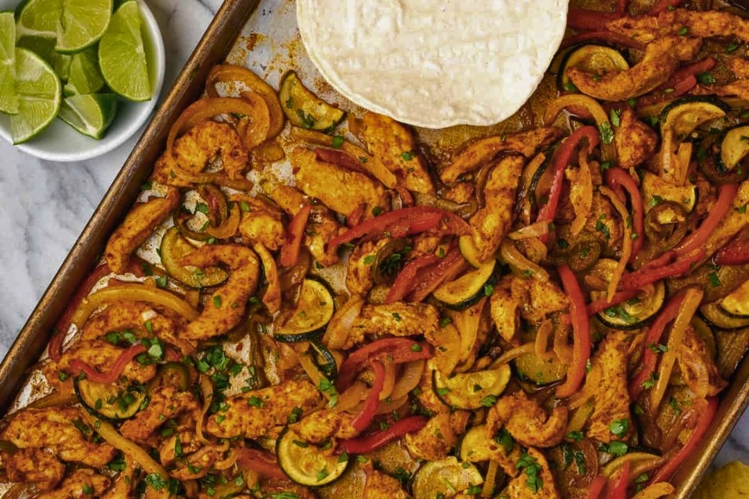 Sheet pan with cooked chicken strips, sliced red peppers, zucchini, and onions, next to a bowl of lime wedges and a stack of tortillas—perfect for easy dinners or flavorful weeknight meals.