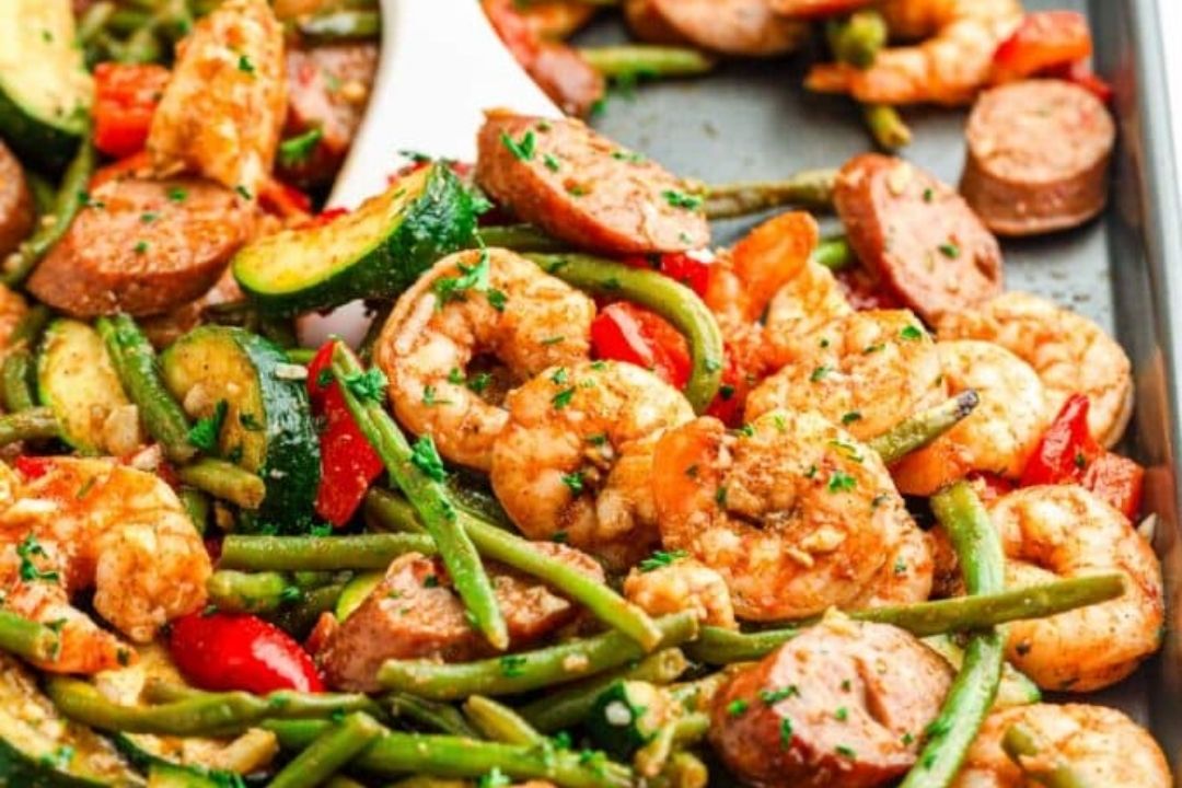 Sheet pan with cooked shrimp, sliced sausage, green beans, zucchini, and red bell peppers, garnished with herbs—perfect for easy recipes and effortless weeknight dinners.