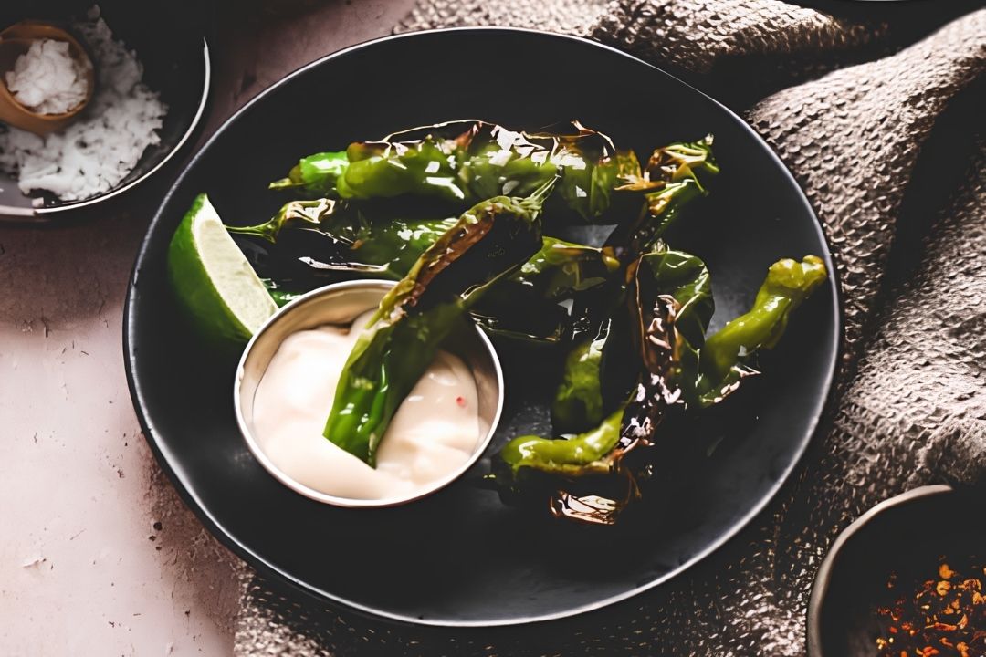 A black plate with roasted green peppers, a lime wedge, and a small metal cup of creamy dipping sauce on a textured cloth—perfect inspiration for easy vegetable dishes or new vegetarian recipes.