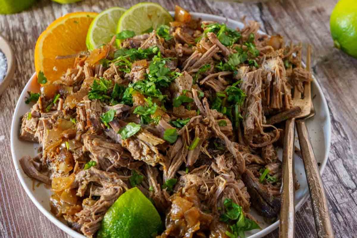 A plate of shredded meat garnished with chopped herbs, served with lime and orange slices on a wooden table, evokes the comforting flavors of fall recipes. Two forks rest beside the plate, making it perfect for sharing a delicious slow cooker meal.