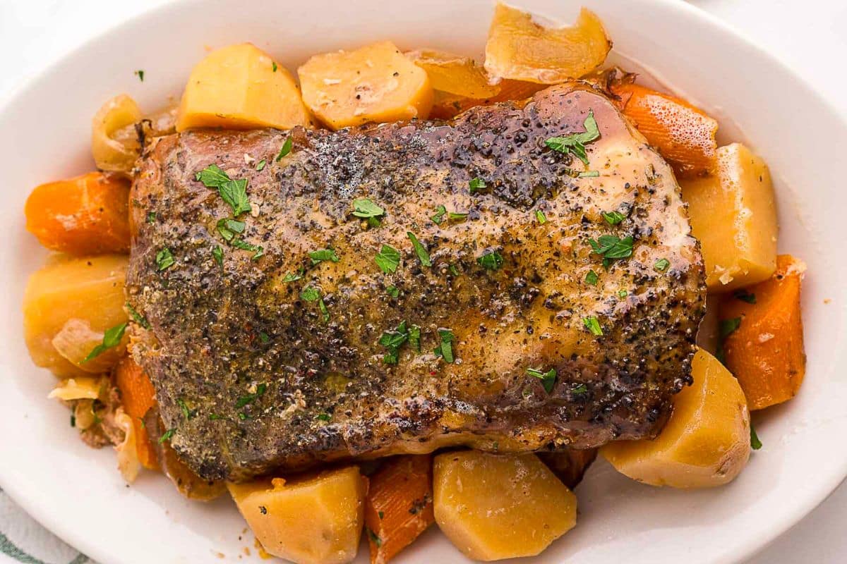 A seasoned pork roast served on a white dish, surrounded by chunks of carrots and potatoes, garnished with chopped herbs—this is one of those classic slow cooker fall recipes that fills your home with comforting aromas all day long.
