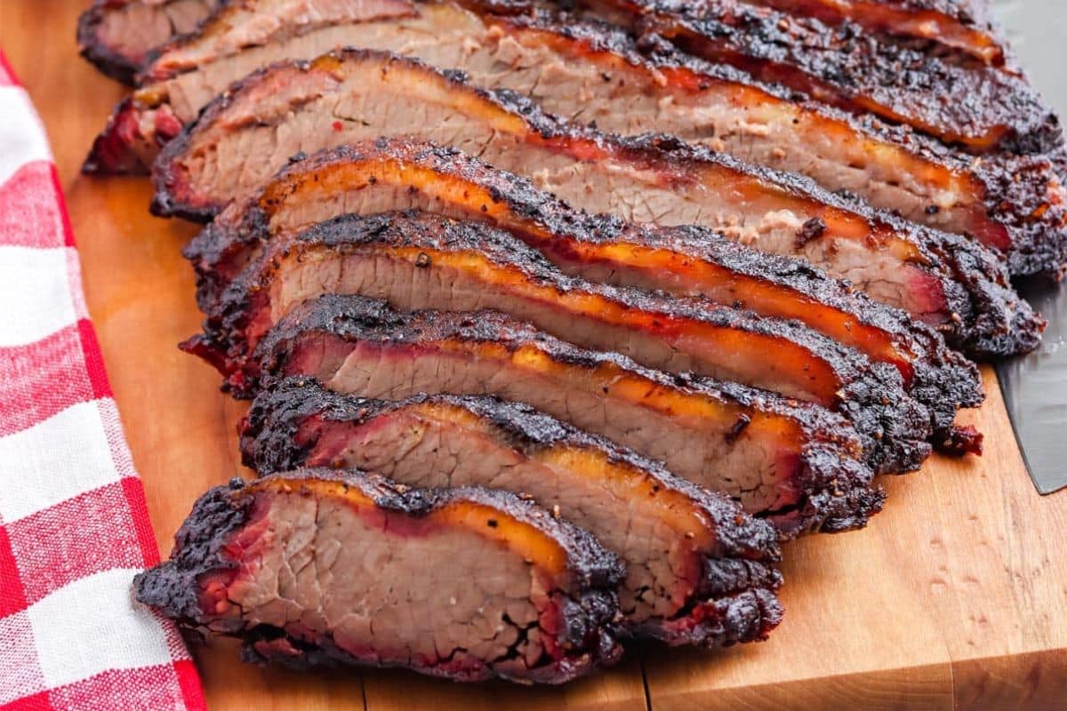 Sliced smoked brisket with a dark bark and visible smoke ring, arranged on a wooden cutting board next to a red and white checkered cloth—perfect for Labor Day cookout recipes or any of your favorite summer recipes.