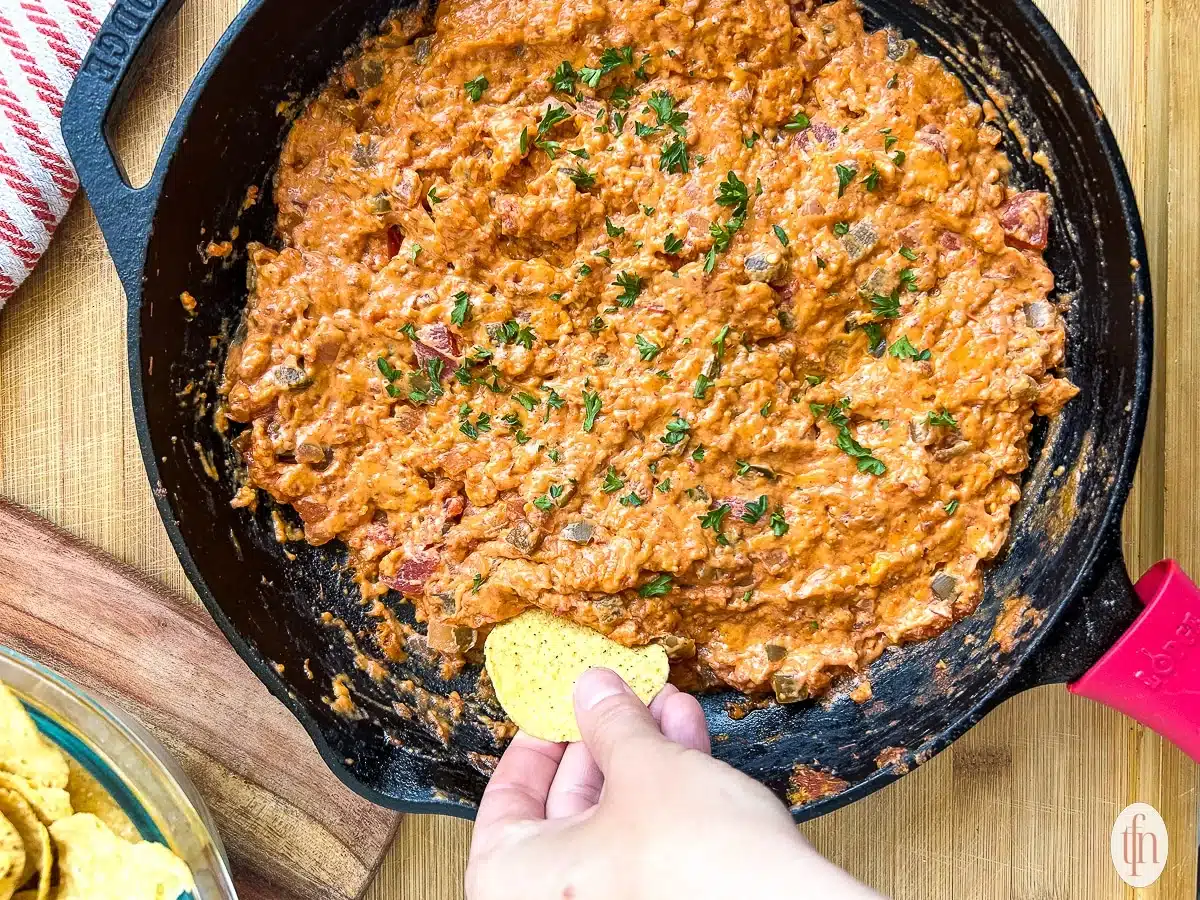 A hand dips a tortilla chip into a cast iron skillet filled with cheesy, tomato-based dip, garnished with fresh herbs—one of those perfect Labor Day cookout recipes. Other chips are visible in a glass dish on the side, inviting everyone to dig into this crowd-pleasing summer recipe.