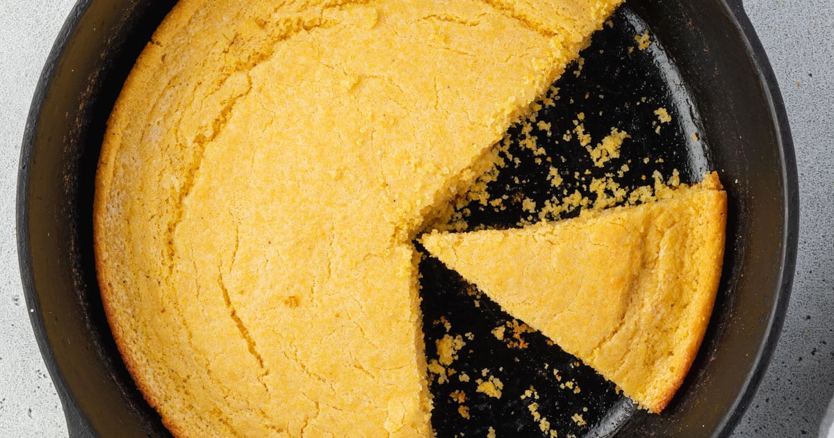 A round cornbread in a black skillet with one triangular slice removed and a few crumbs left behind—perfect as a make-ahead addition to your Labor Day sides for stress-free hosting.
