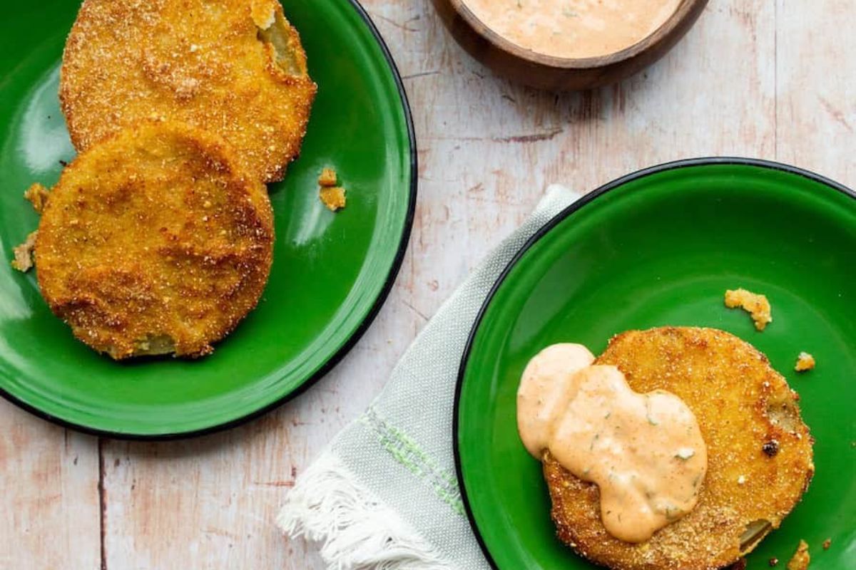 Two green plates with fried green tomato slices—a classic among summer recipes and a favorite in garden tomato recipes—are arranged on a light wooden surface. One slice is topped with a creamy sauce, while a small bowl of sauce and a napkin complete this inviting display of fresh tomato dishes.