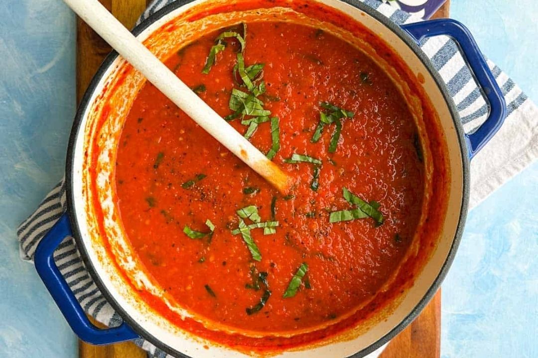 A pot of tomato sauce garnished with chopped basil sits on a striped towel, with a wooden spoon resting inside.