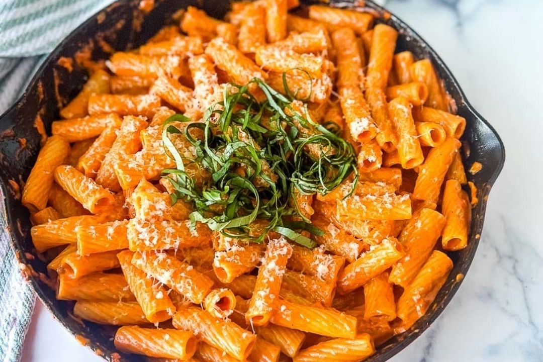A skillet filled with rigatoni pasta in a creamy tomato sauce, topped with grated cheese and fresh chopped basil—perfect for those seeking cozy pasta dinners.