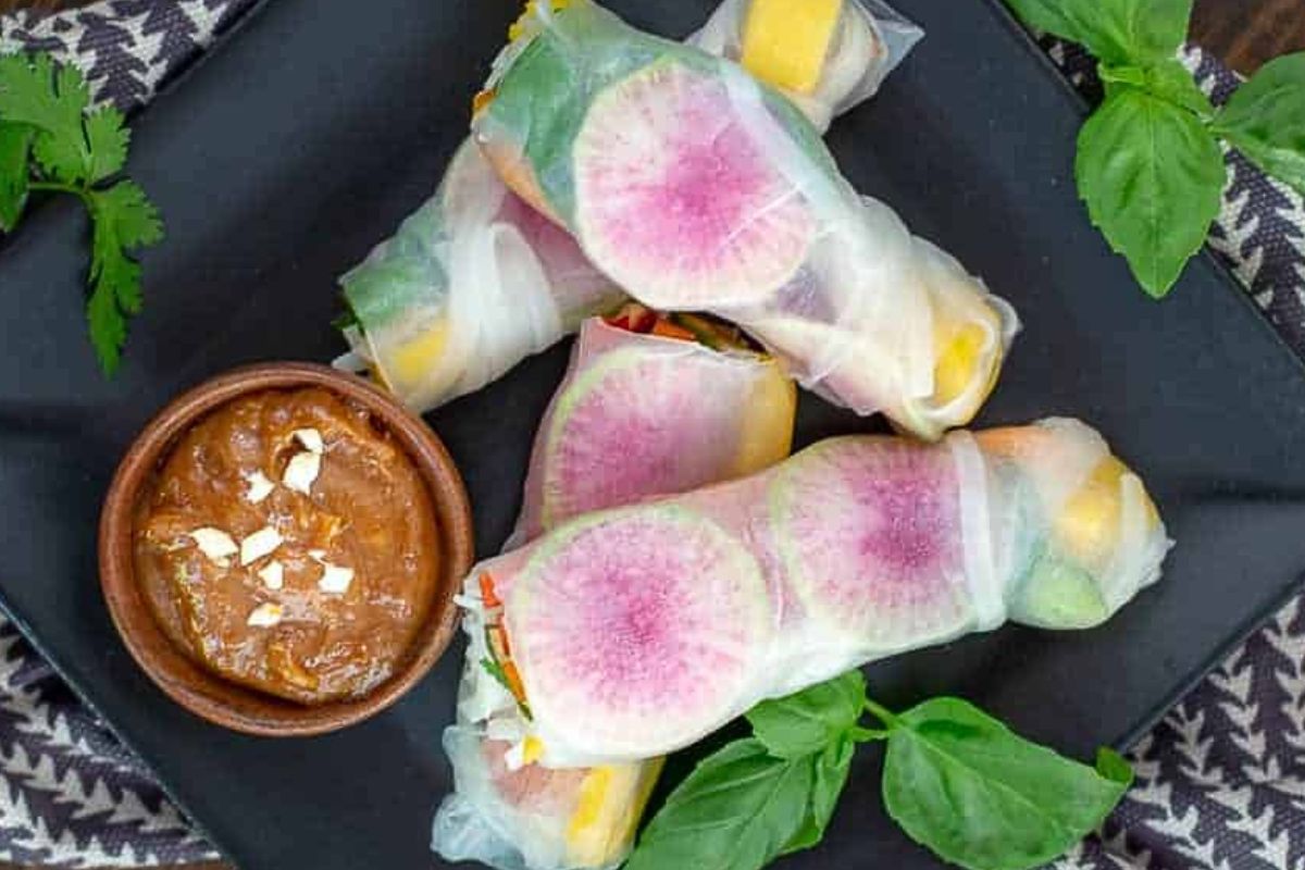 Three fresh spring rolls with visible radish slices are arranged on a black plate with a small bowl of dipping sauce, garnished with herbs on the side—a perfect addition to your collection of unique luncheon ideas.