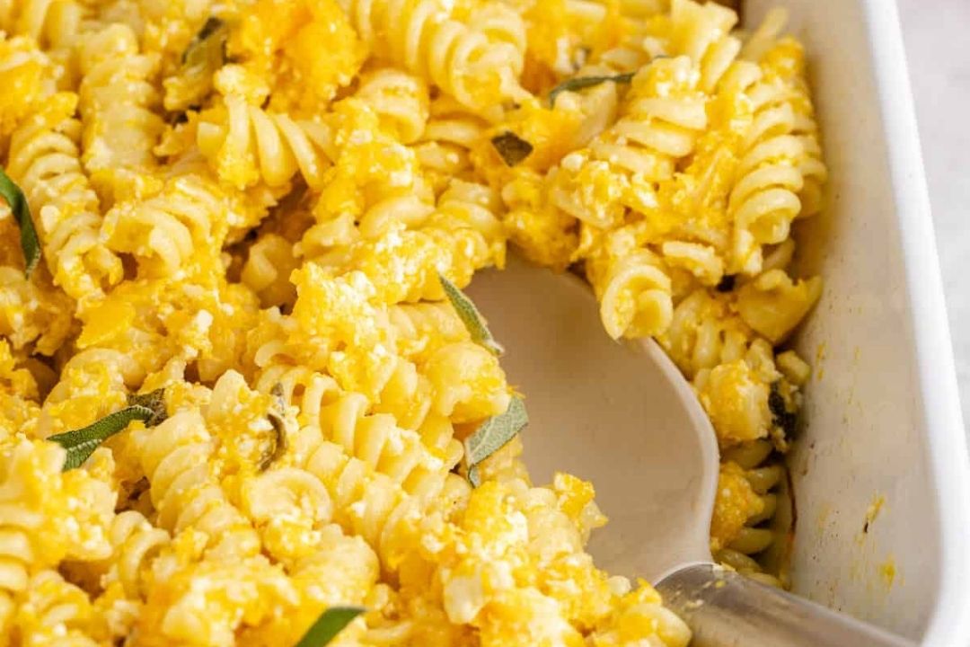 Baked rotini pasta with a creamy yellow sauce and fresh herb pieces—perfect for fall recipes—served in a white dish with a metal serving spoon.