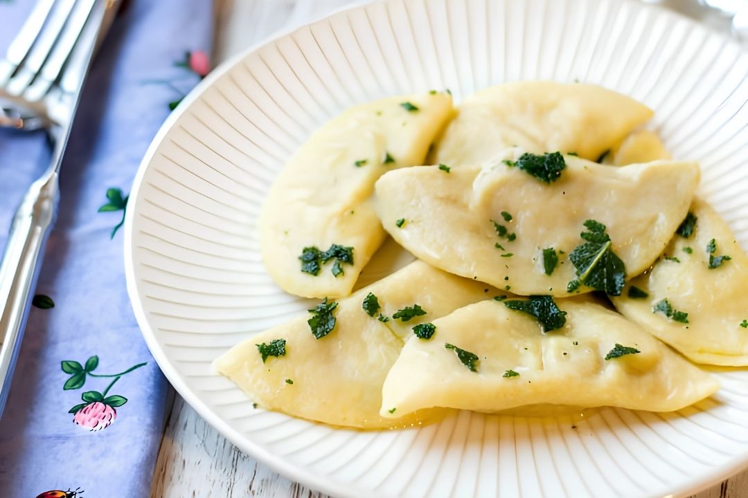 A plate of six folded dumplings, perfect for autumn meals, is garnished with chopped herbs and sits on a white patterned dish beside a fork and a floral napkin.