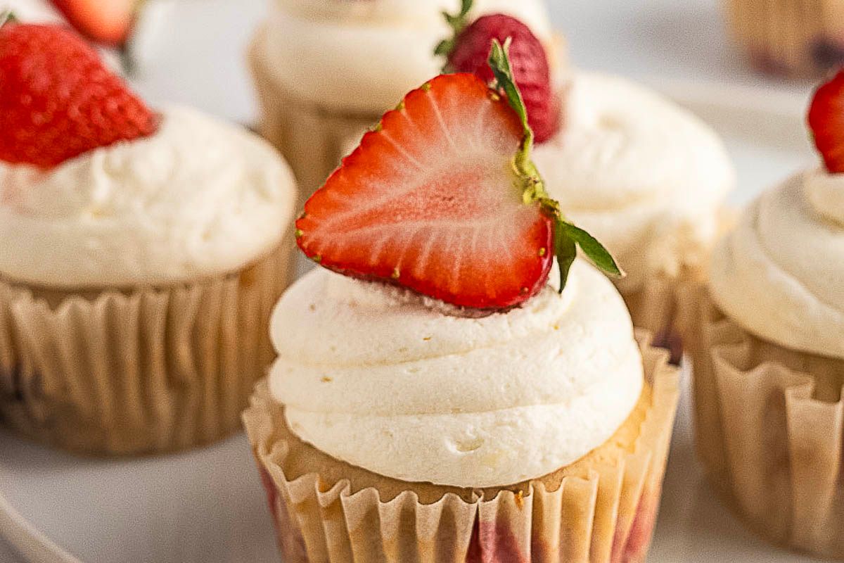 Gluten free vanilla cupcakes with swirled white frosting, each topped with a halved strawberry, sit on a white surface—making them the perfect treat for summer desserts.