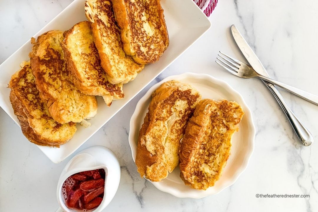 A rectangular plate and a small round plate with slices of French toast, a bowl of strawberries in syrup, and a fork and knife on a marble surface.