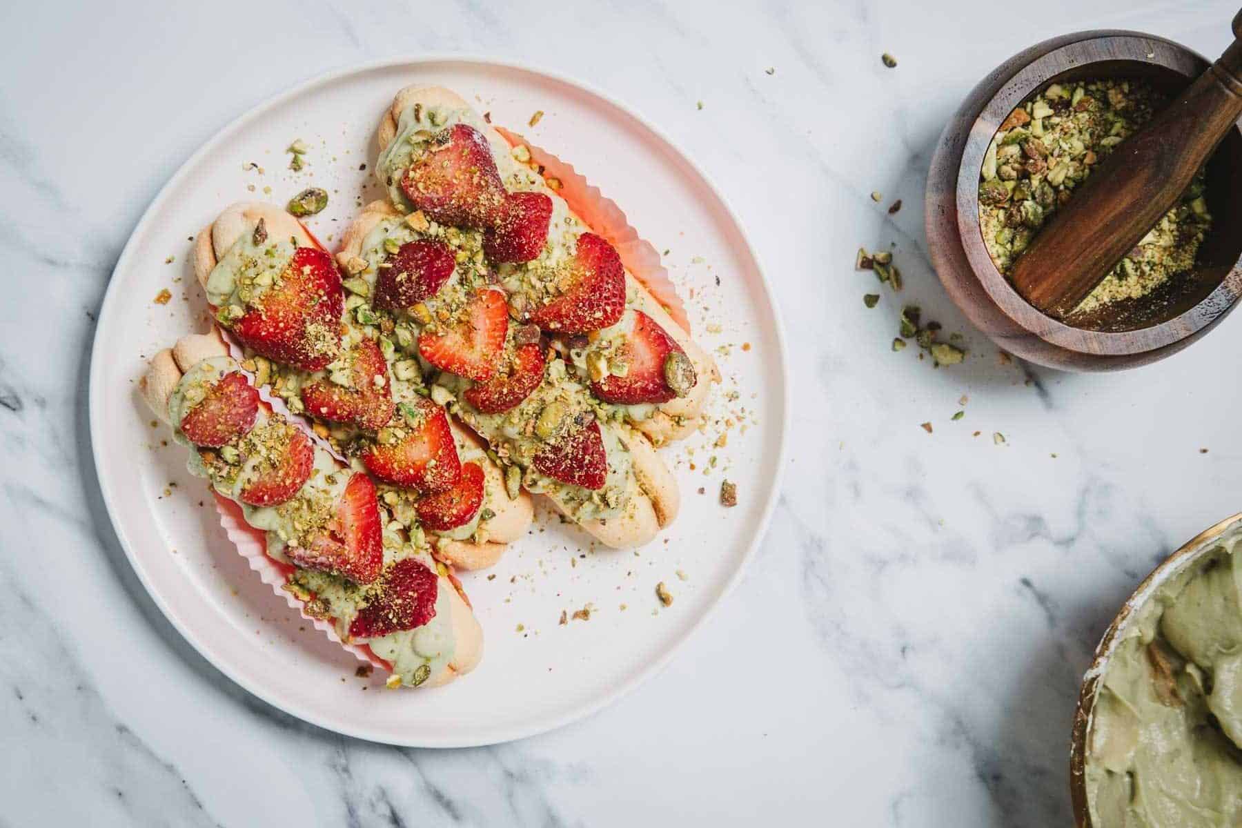 A plate of ladyfingers topped with cream, sliced strawberries, and crushed pistachios—perfect for fresh recipes—sits beside a wooden bowl of ground pistachios and a pestle on a marble surface.