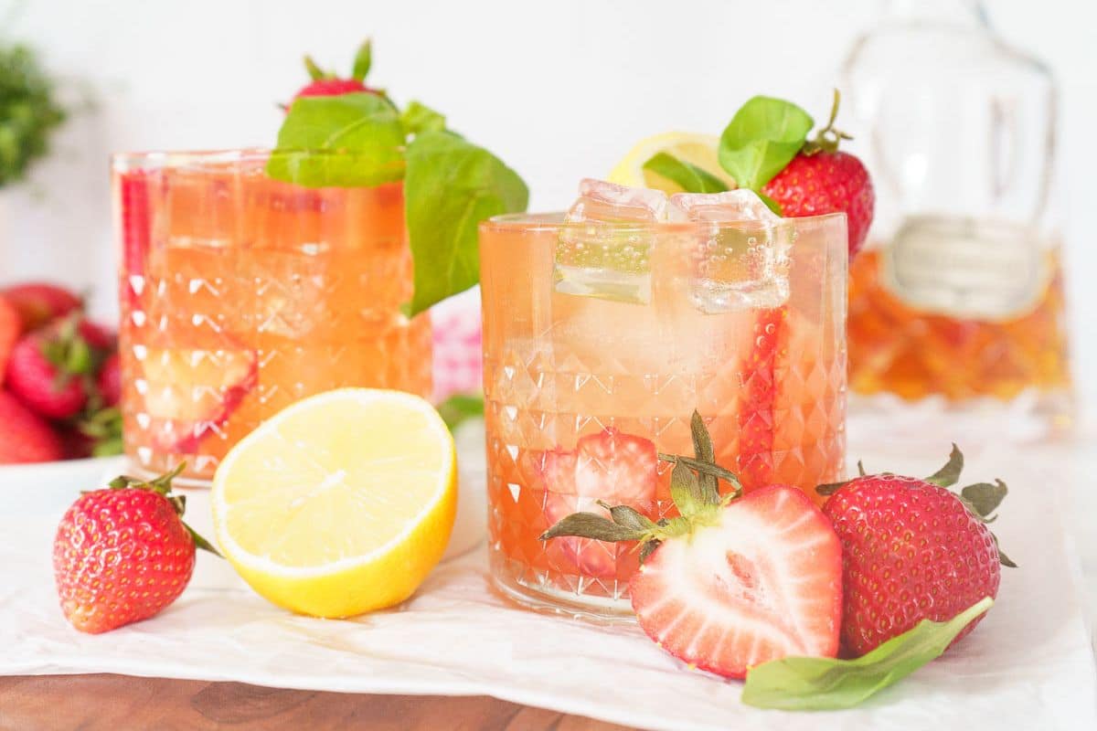 Two glasses of pink iced cocktails garnished with fresh strawberries, basil leaves, and lemon make the perfect summer refreshments. Surrounded by whole and halved strawberries and a lemon half on a white surface, these cool drinks are ideal for your Labor Day beverages spread.