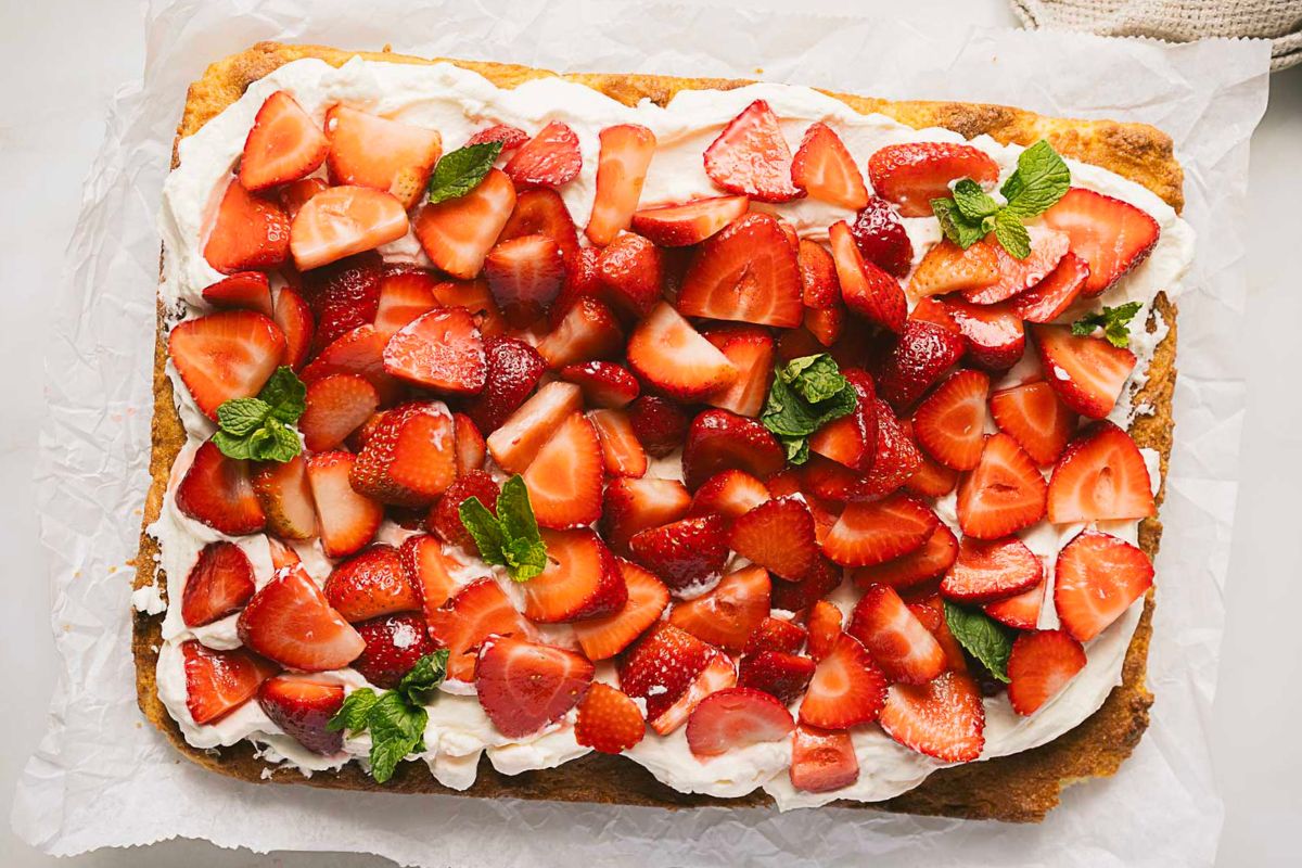 Rectangular cake topped with whipped cream, sliced fresh strawberries, and mint leaves, displayed on parchment paper—perfect for showcasing your favorite Gluten-Free Recipes at summer gatherings or as a festive treat for Labor Day Recipes.