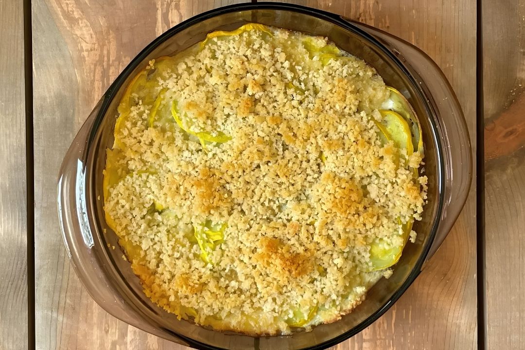 A glass baking dish filled with baked yellow squash casserole topped with a golden, crumbly breadcrumb layer, placed on a wooden surface.