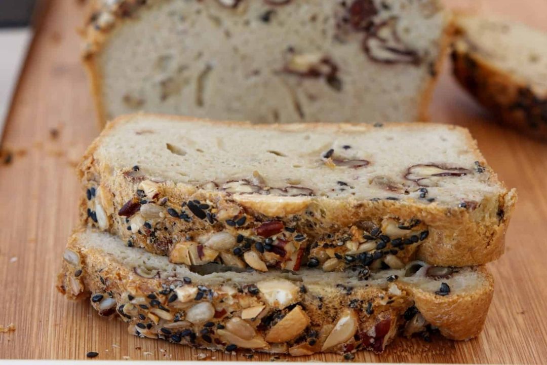 Two slices of seeded bread with nuts and seeds visible on the crust and inside, placed on a wooden cutting board with a loaf in the background.