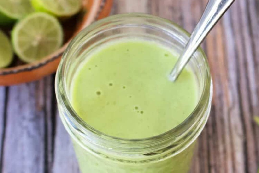 A glass jar filled with creamy green dressing, with a metal spoon inside. Sliced limes are visible in a bowl in the background on a wooden surface.
