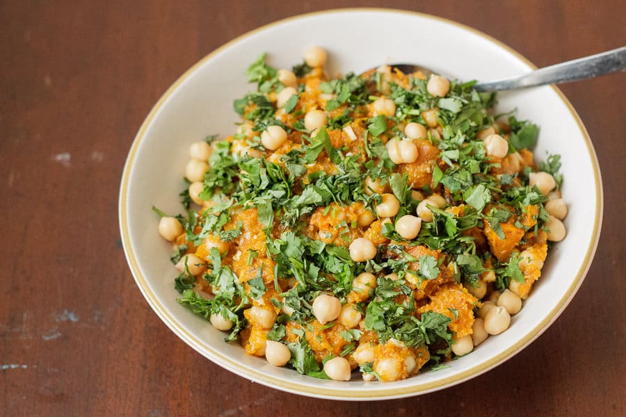 A bowl of chickpea salad with chopped cilantro and orange vegetables, possibly sweet potatoes or squash, on a wooden table with a spoon—an inviting addition to your collection of comforting autumn meals.