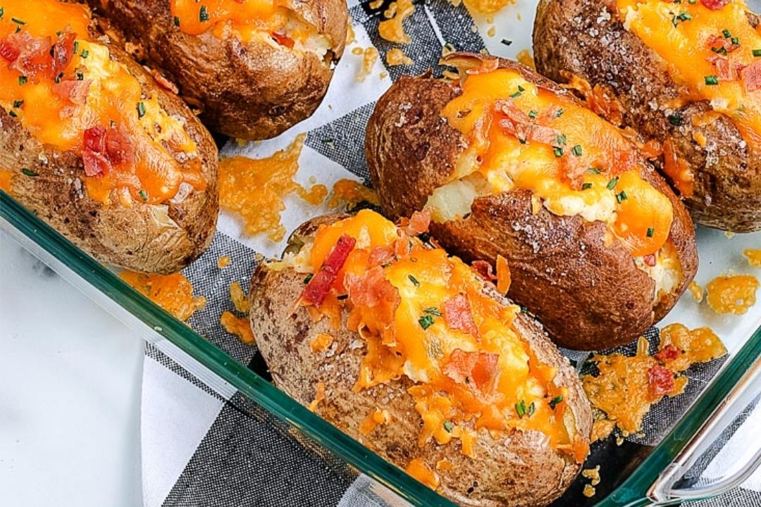 Baked potatoes in a glass dish topped with melted cheddar cheese, bacon bits, and chives on a checkered cloth background.