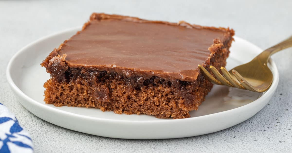 A single square piece of chocolate sheet cake with chocolate icing sits on a white plate alongside a gold fork—an irresistible treat that’s sure to become one of your tried and true recipes for popular potluck dishes.