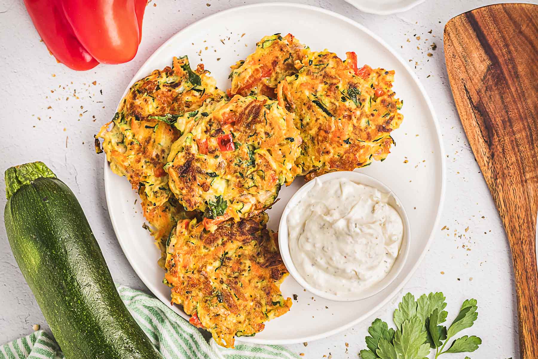 A plate of zucchini fritters with red peppers, served with a small bowl of dipping sauce; zucchini, parsley, and a red pepper are nearby—one of our reader favorites among popular side dishes.