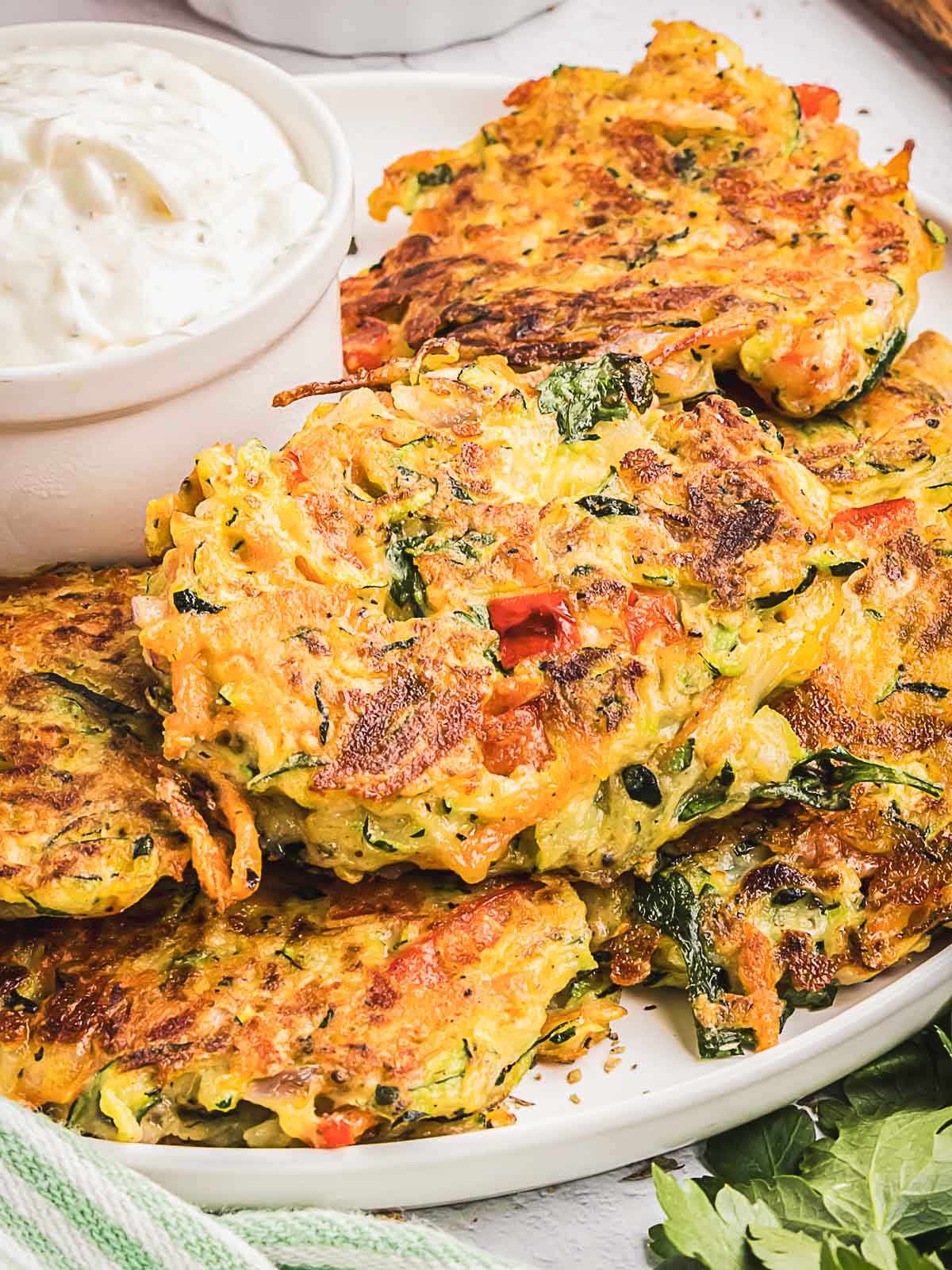 A plate of crispy vegetable fritters with visible greens and red peppers, served alongside a bowl of creamy garlic sauce.