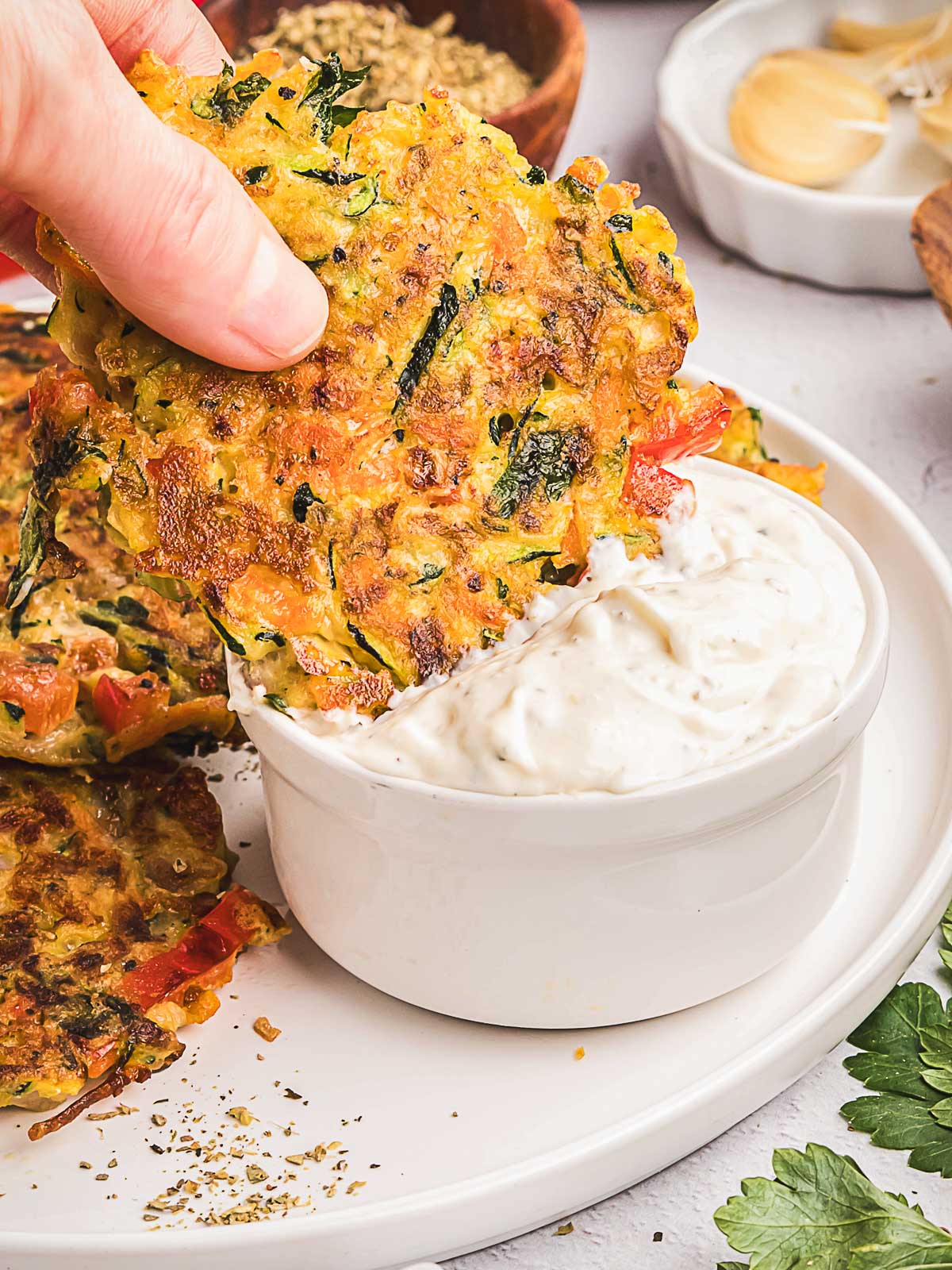 A hand dips a crispy vegetable fritter into a bowl of creamy garlic sauce on a plate with more crispy fritters and fresh herbs nearby.