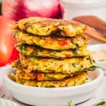 A stack of golden-brown crispy fritters sits on a white plate, with visible pieces of zucchini, red pepper, and herbs.