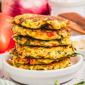A stack of golden-brown crispy fritters sits on a white plate, with visible pieces of zucchini, red pepper, and herbs.