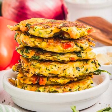 A stack of golden-brown crispy fritters sits on a white plate, with visible pieces of zucchini, red pepper, and herbs.