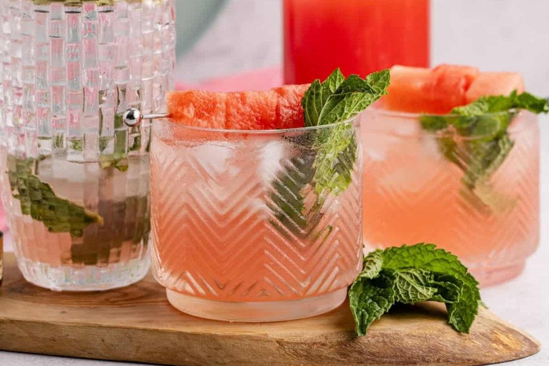 Three glasses of pink watermelon cocktails with ice, mint leaves, and watermelon slices are arranged on a wooden board—perfect summer refreshments to enjoy on a warm afternoon.