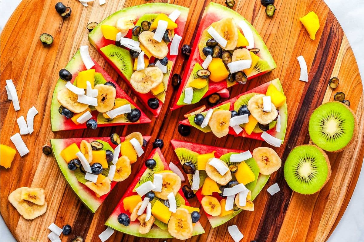 Sliced watermelon topped with banana, blueberries, mango, coconut flakes, and dried banana chips, arranged on a wooden board with kiwi halves and extra toppings scattered around—this colorful spread is perfect for easy desserts at cookouts or festive Labor Day gatherings.