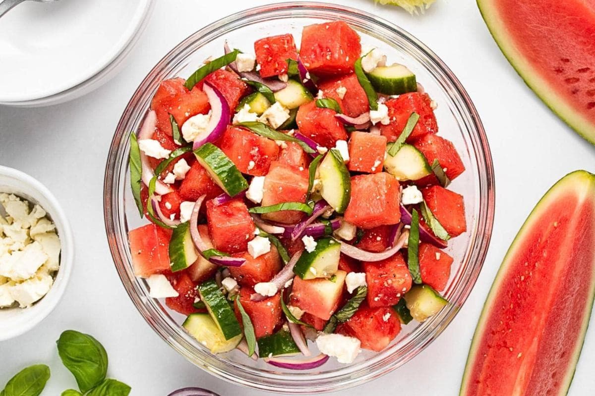 A glass bowl filled with watermelon, cucumber, red onion, feta cheese, and fresh basil salad—a perfect addition to your summer recipes. Sliced ingredients and bowls of feta and salt are nearby, making this dish a celebration of garden-fresh recipes for seasonal cooking.