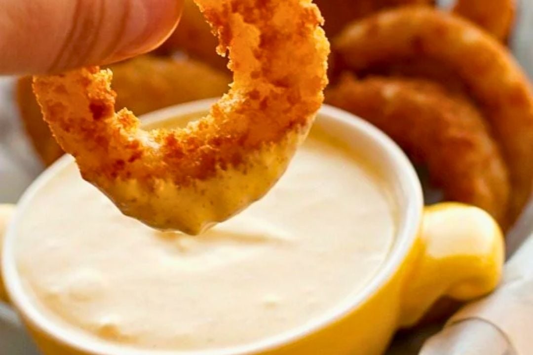 A hand dips a crispy onion ring into a bowl of creamy cold dip, with more onion rings visible in the background—perfect for easy snacking on game day.