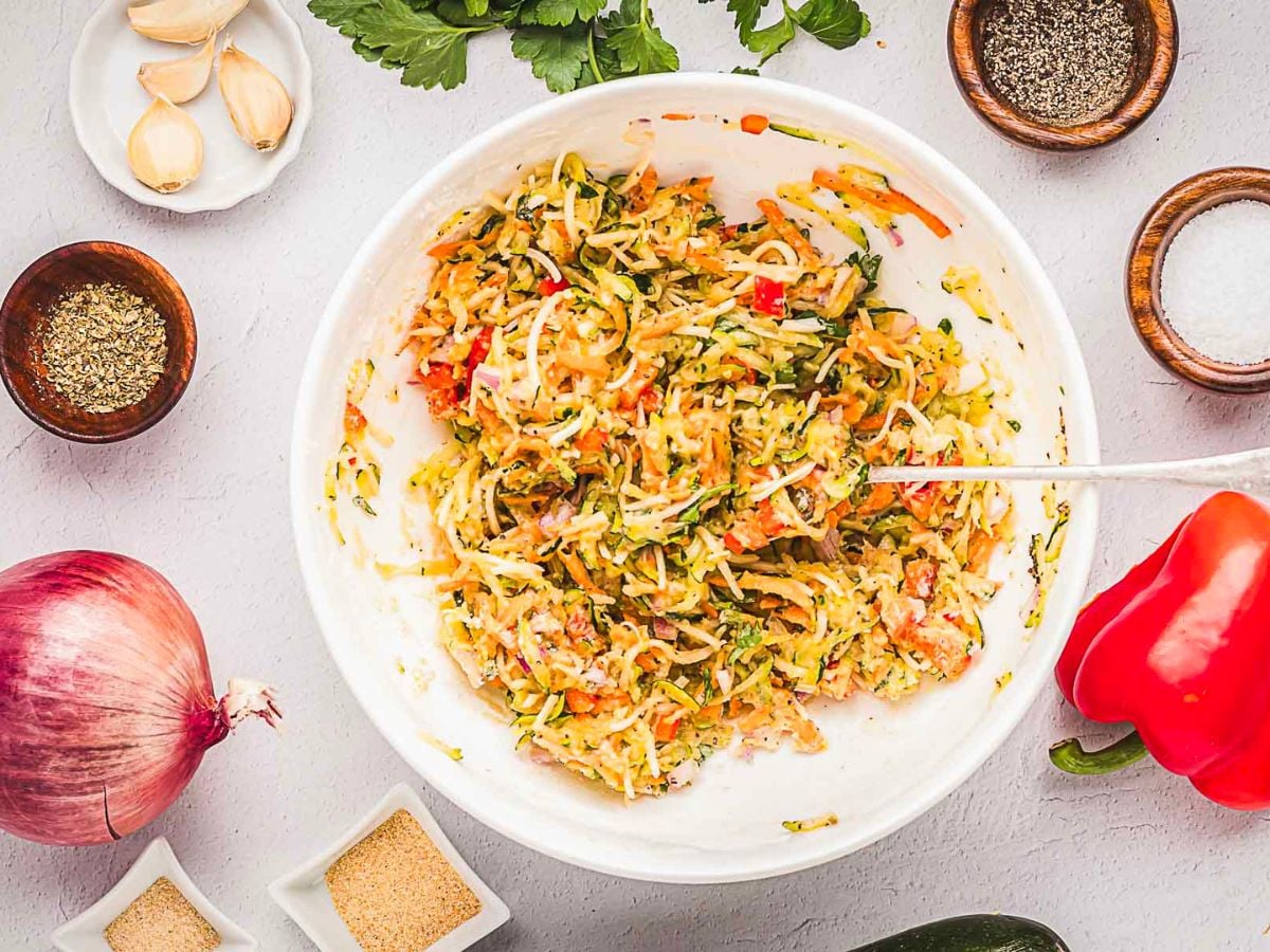 A white bowl filled with a mixed vegetable batter for crispy vegetable fritters, surrounded by raw garlic, onion, red bell pepper, spices, and fresh herbs on a light surface-perfect for pairing with a zesty garlic sauce.