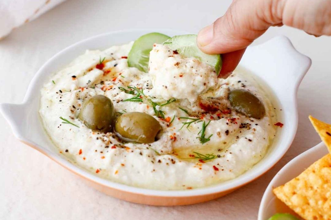 A hand dips a cucumber slice into a bowl of creamy dip garnished with olives, herbs, and spices.