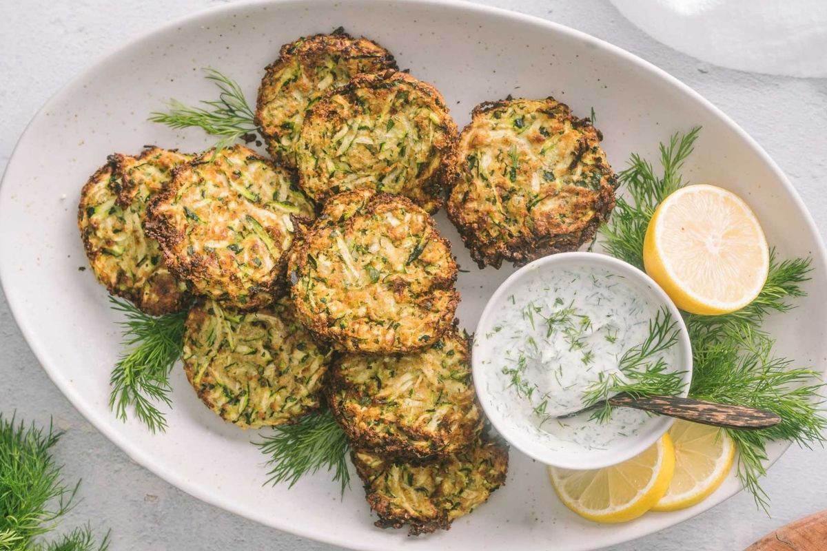 Oval platter with several zucchini fritters, a bowl of creamy dipping sauce with herbs, fresh dill, and lemon slices arranged decoratively on the side—perfect for those seeking delicious vegetarian recipes or creative vegetable dishes.