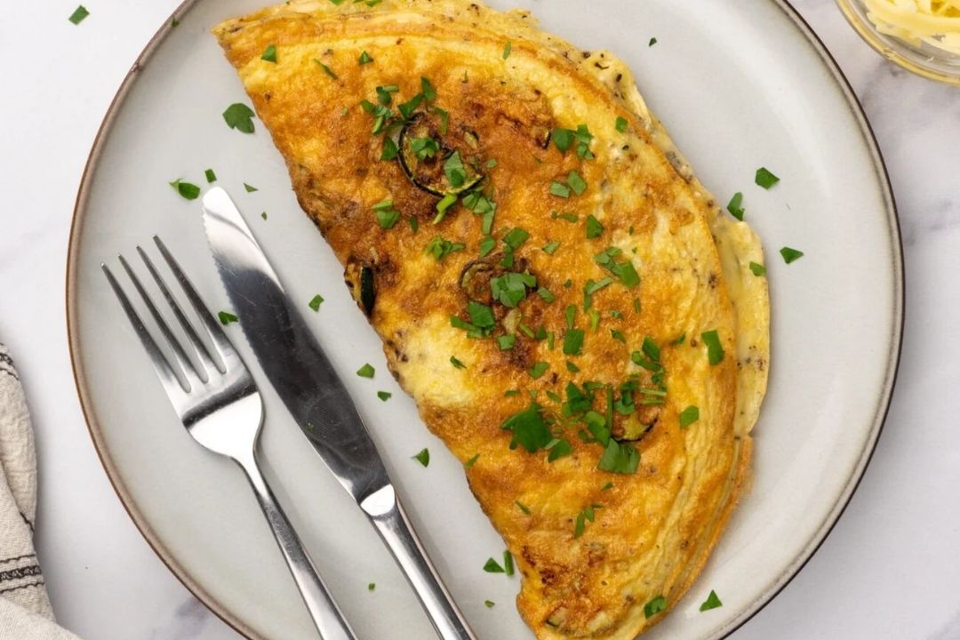 An omelette garnished with chopped parsley is served on a round plate with a fork and knife on the side.